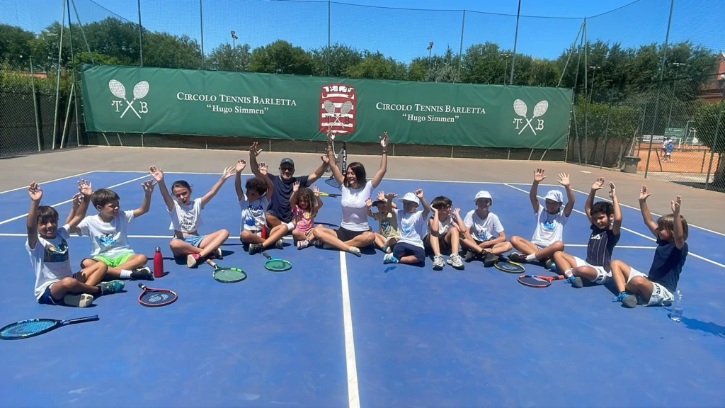Bambini e adulti seduti a gambe incrociate su un campo da tennis, con le braccia alzate.